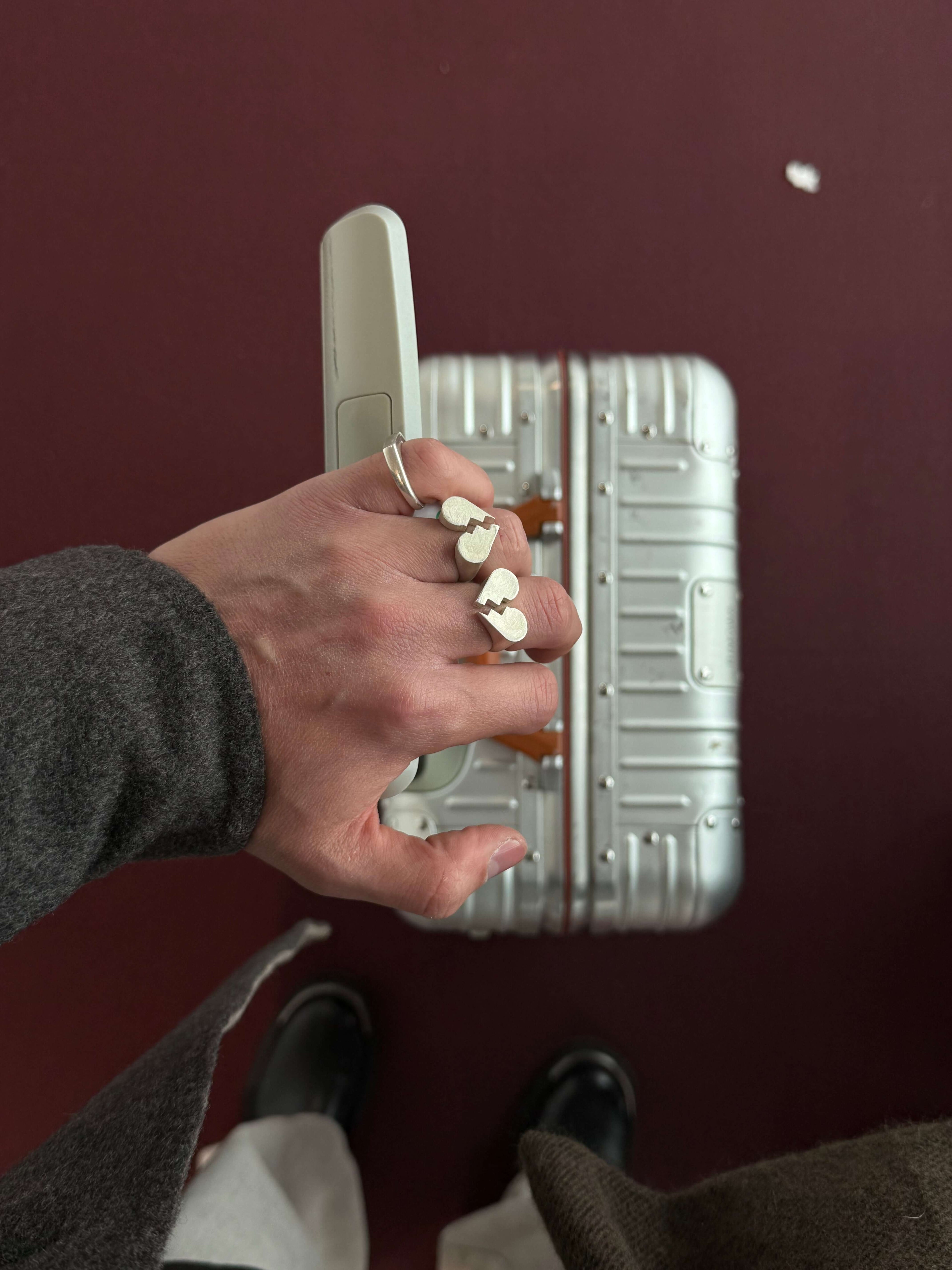 Chunky Jewelry rings adorning a hand holding a silver suitcase against a maroon background.