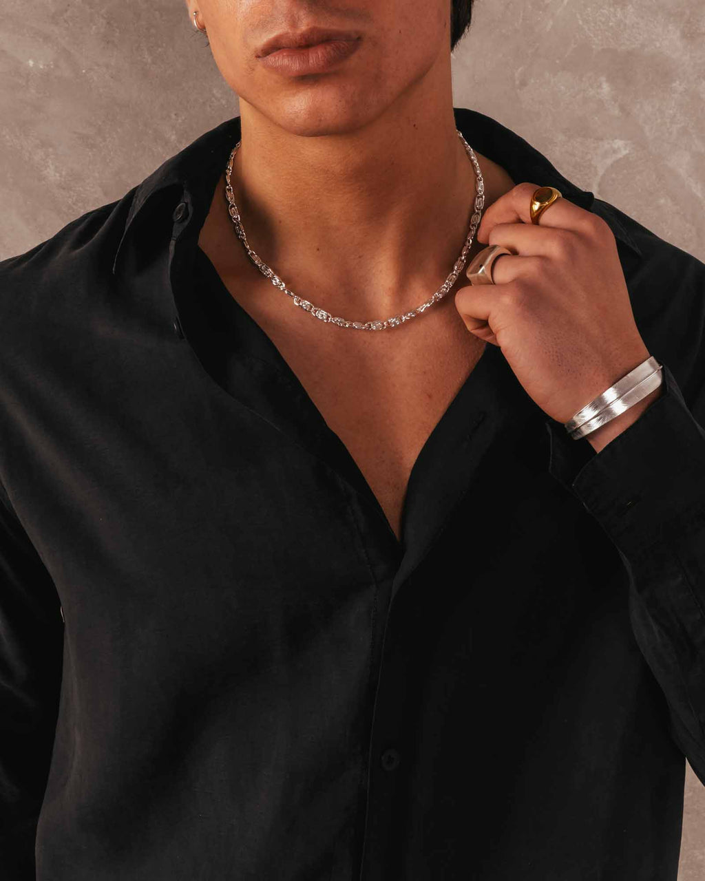 Person wearing a black shirt with jewelry including a necklace, ring, and bracelet against a neutral background.