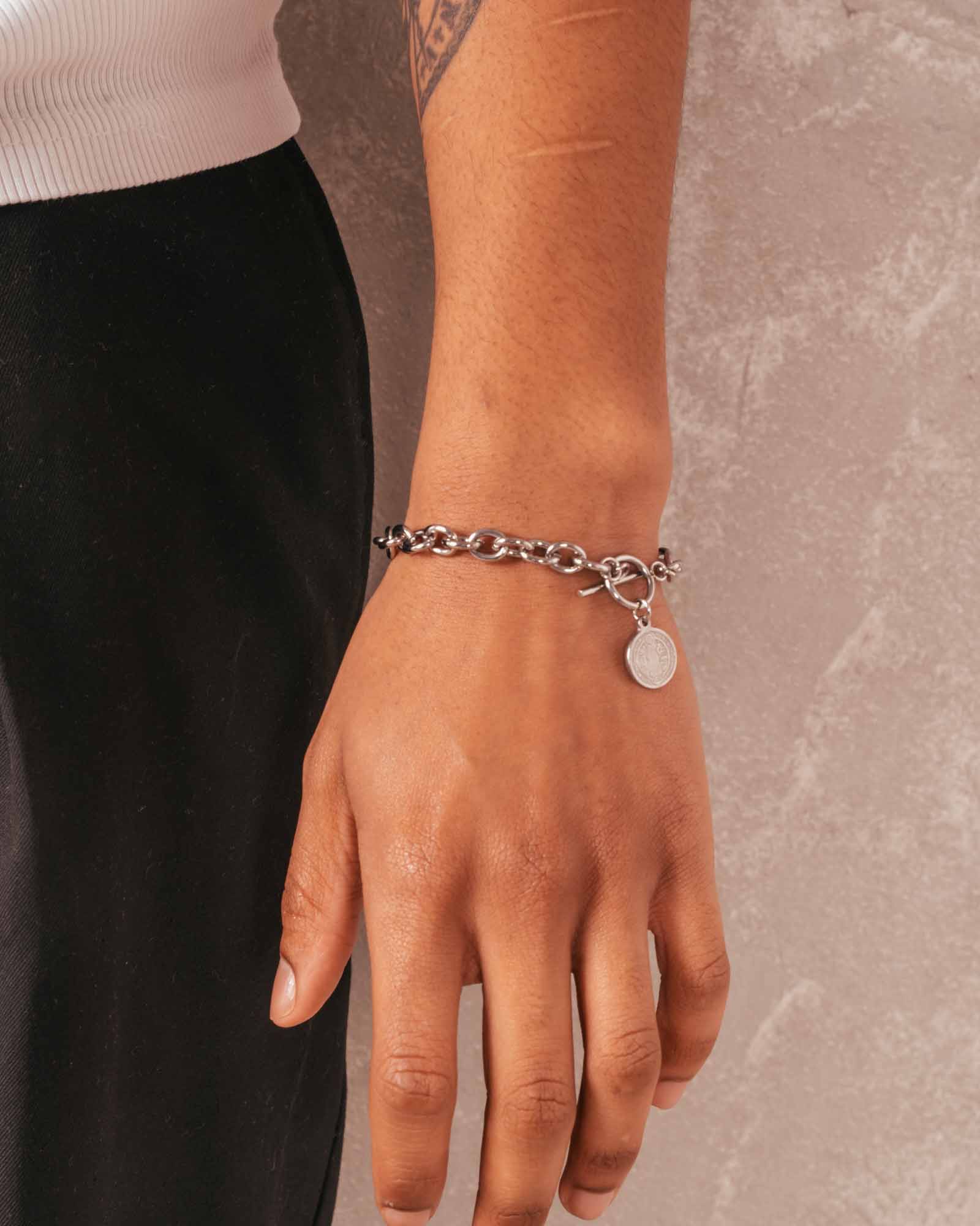 Hand wearing a silver bracelet with a small round tag on a neutral background