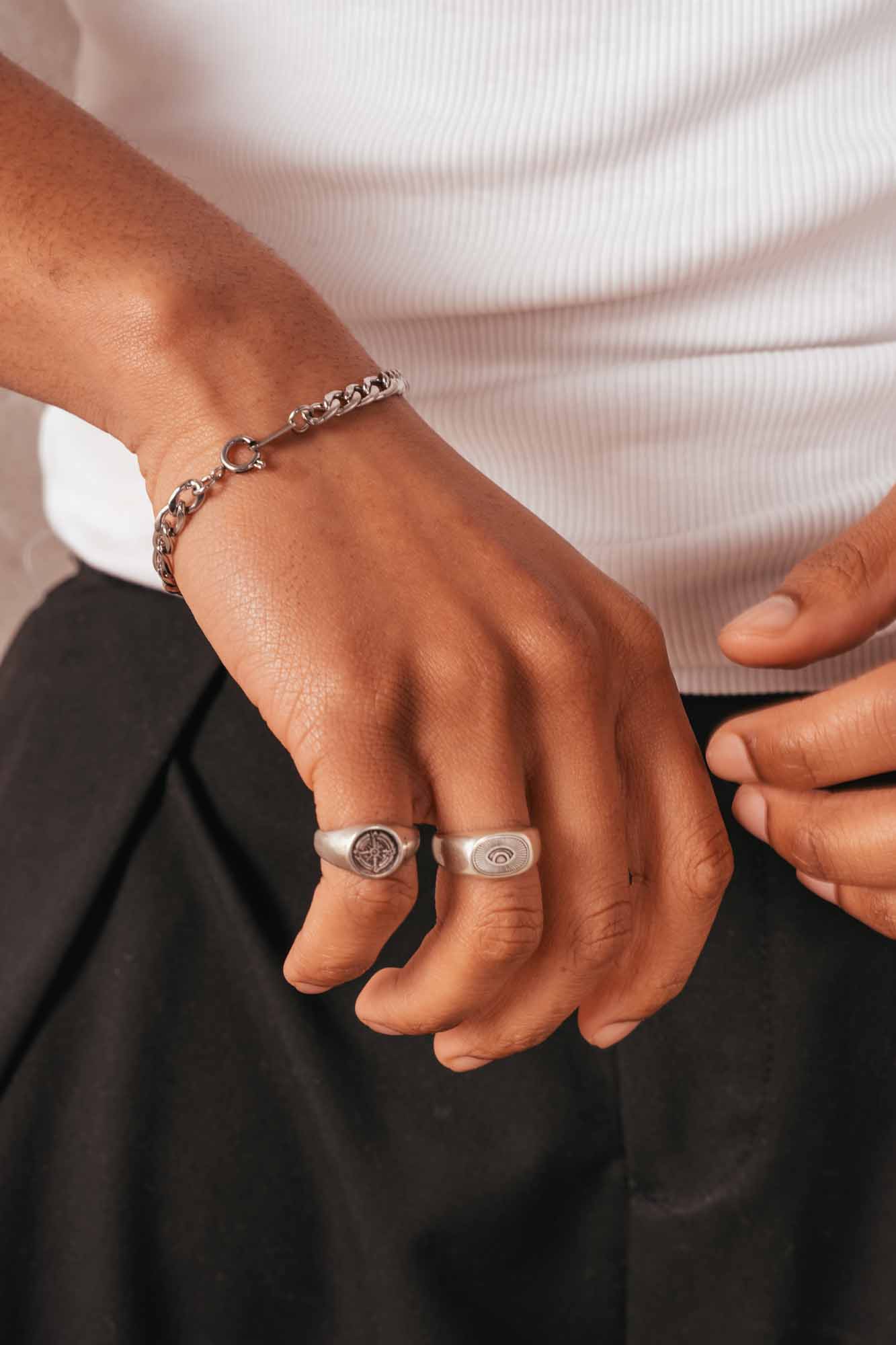 Close-up of a person's wrist with a bracelet and two rings, wearing a white shirt and black pants.