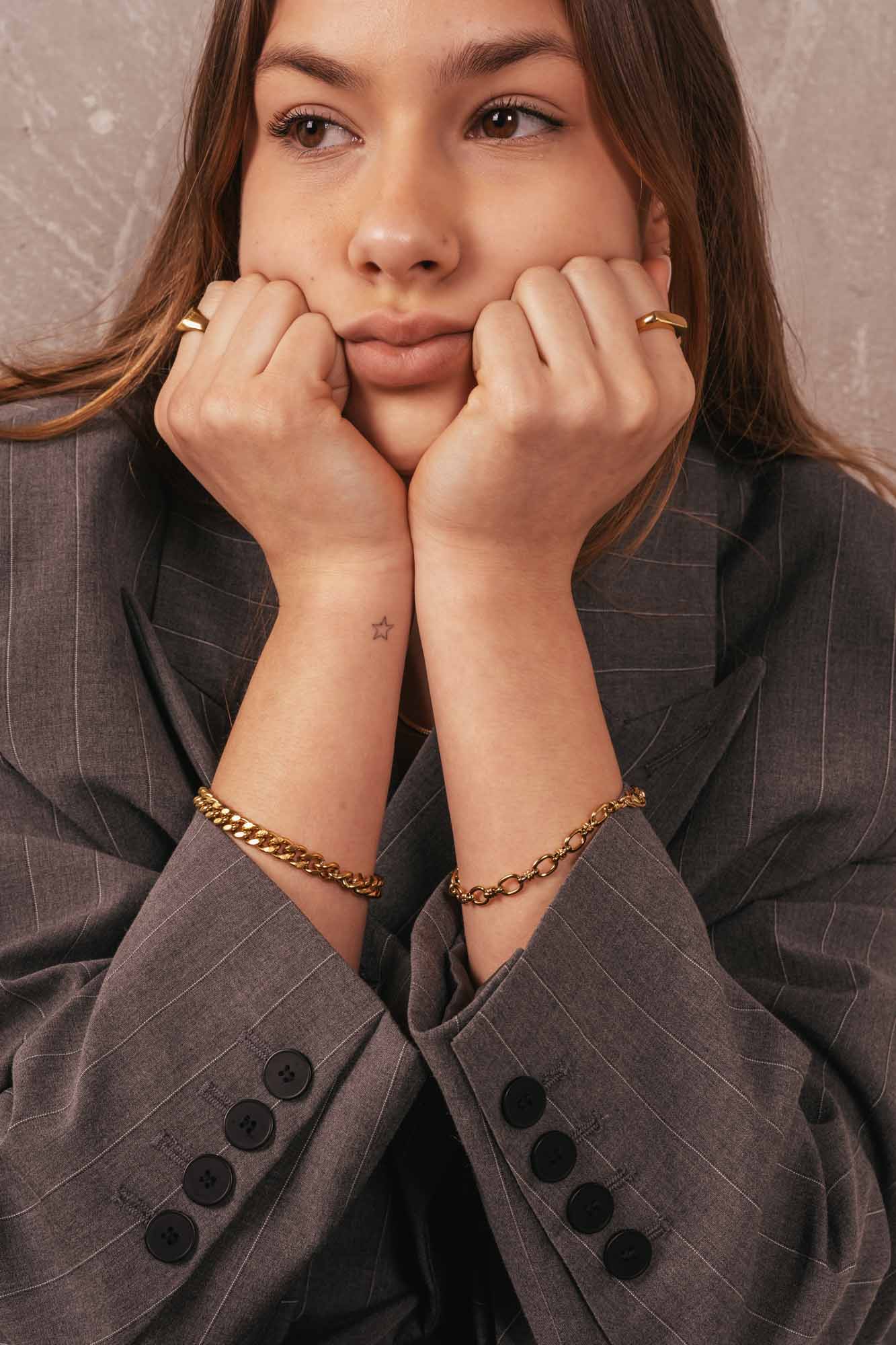 Woman wearing a gray suit with gold jewelry against a neutral background