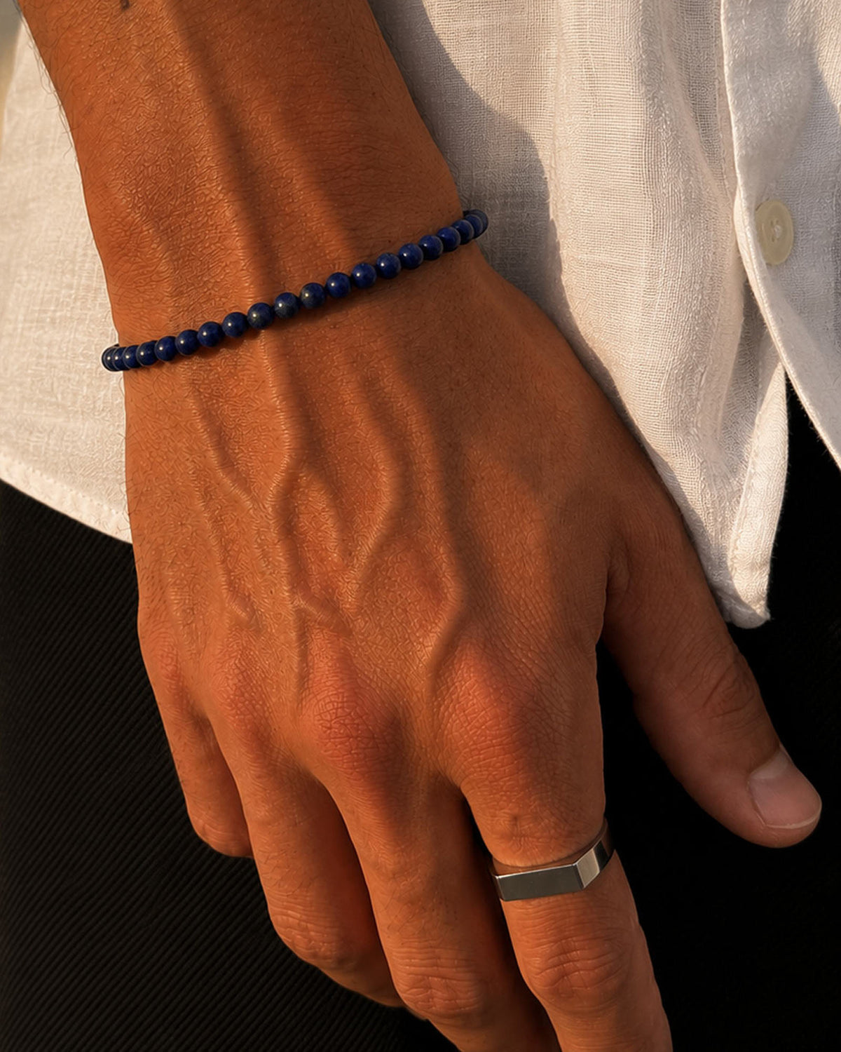 A close-up of a person's hand wearing a dark blue beaded bracelet and a silver ring, with a white shirt in the background.