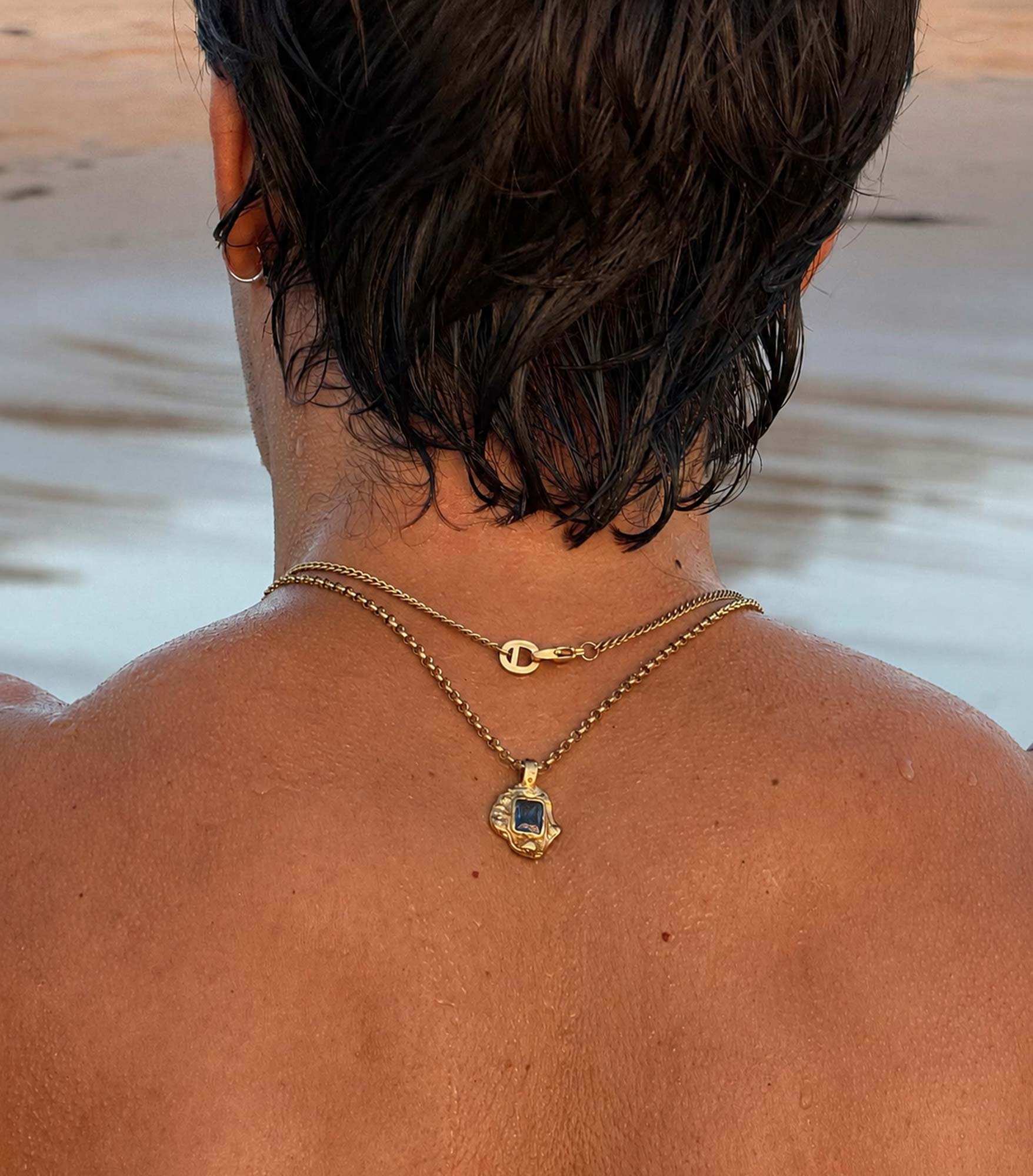 Person wearing a gold necklace with a pendant by a body of water