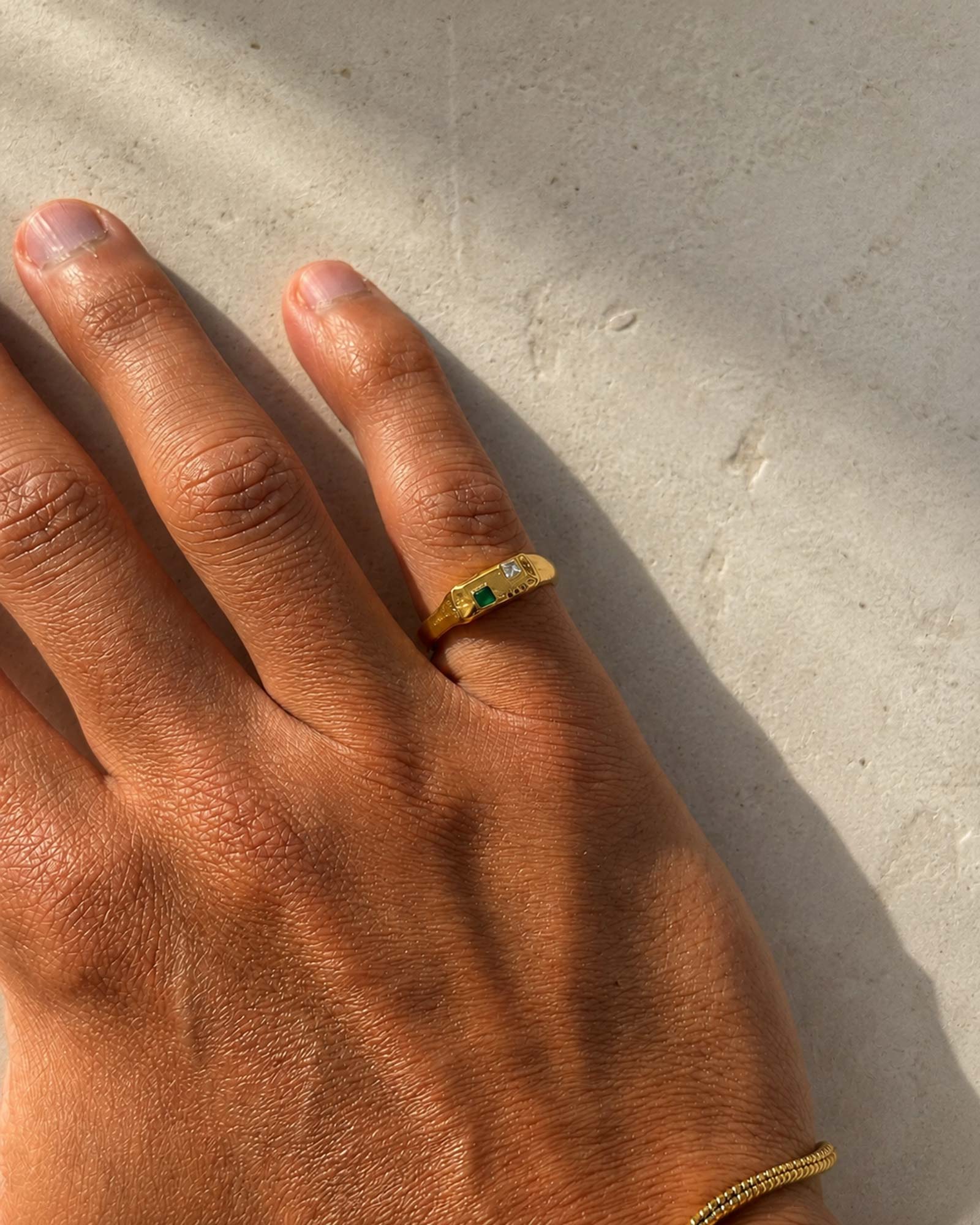 Hand wearing a gold ring with green gemstone on a concrete surface