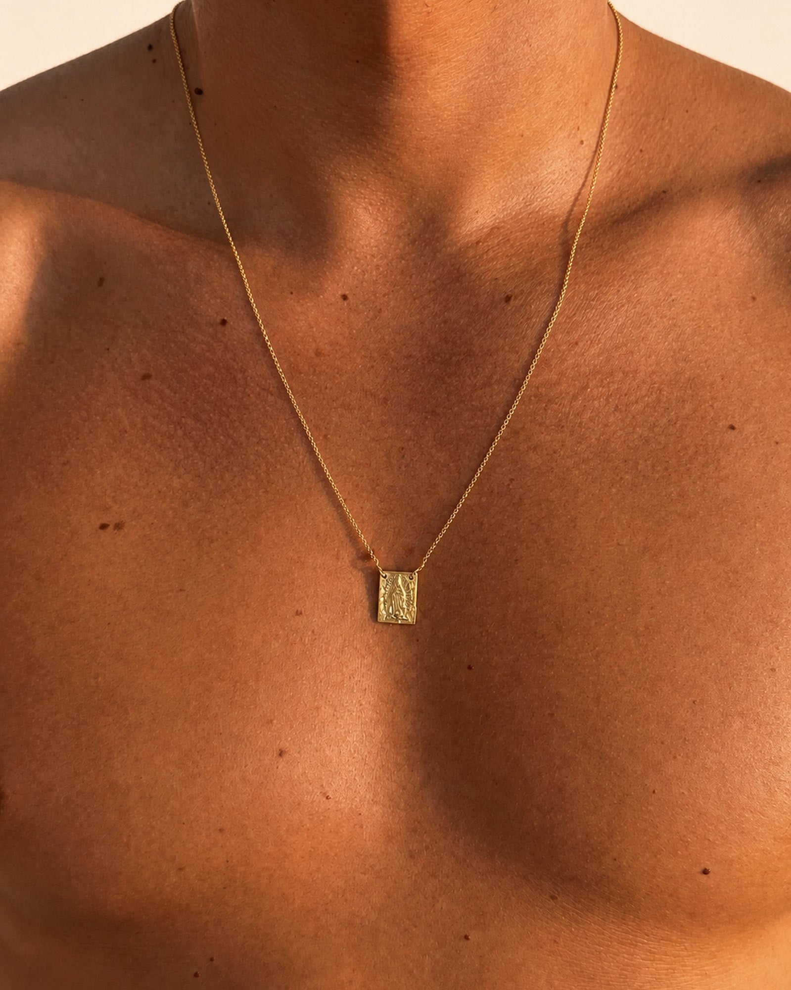 Golden - A close-up of a person's bare chest wearing a delicate gold chain necklace with a small textured rectangular pendant.