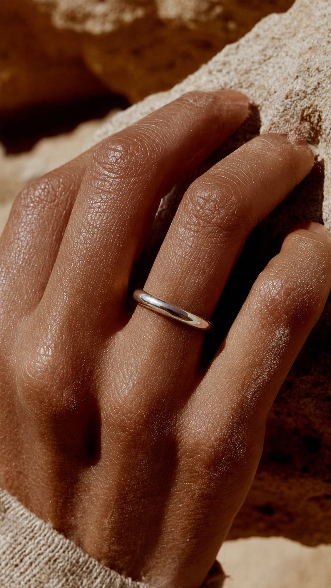 A minimalist, thin silver band ring worn on a person's ring finger, shown in a warm, sunlit desert setting with textured rock and sand in the background.