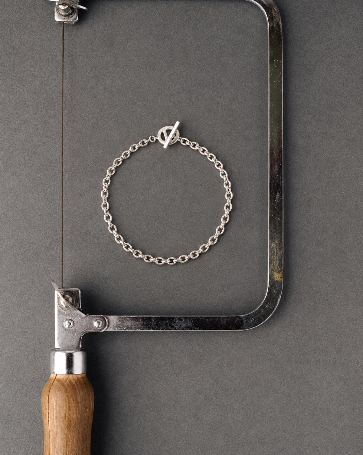 A delicate silver chain bracelet with a small V-shaped clasp, displayed on a dark gray textured background in front of a hacksaw.