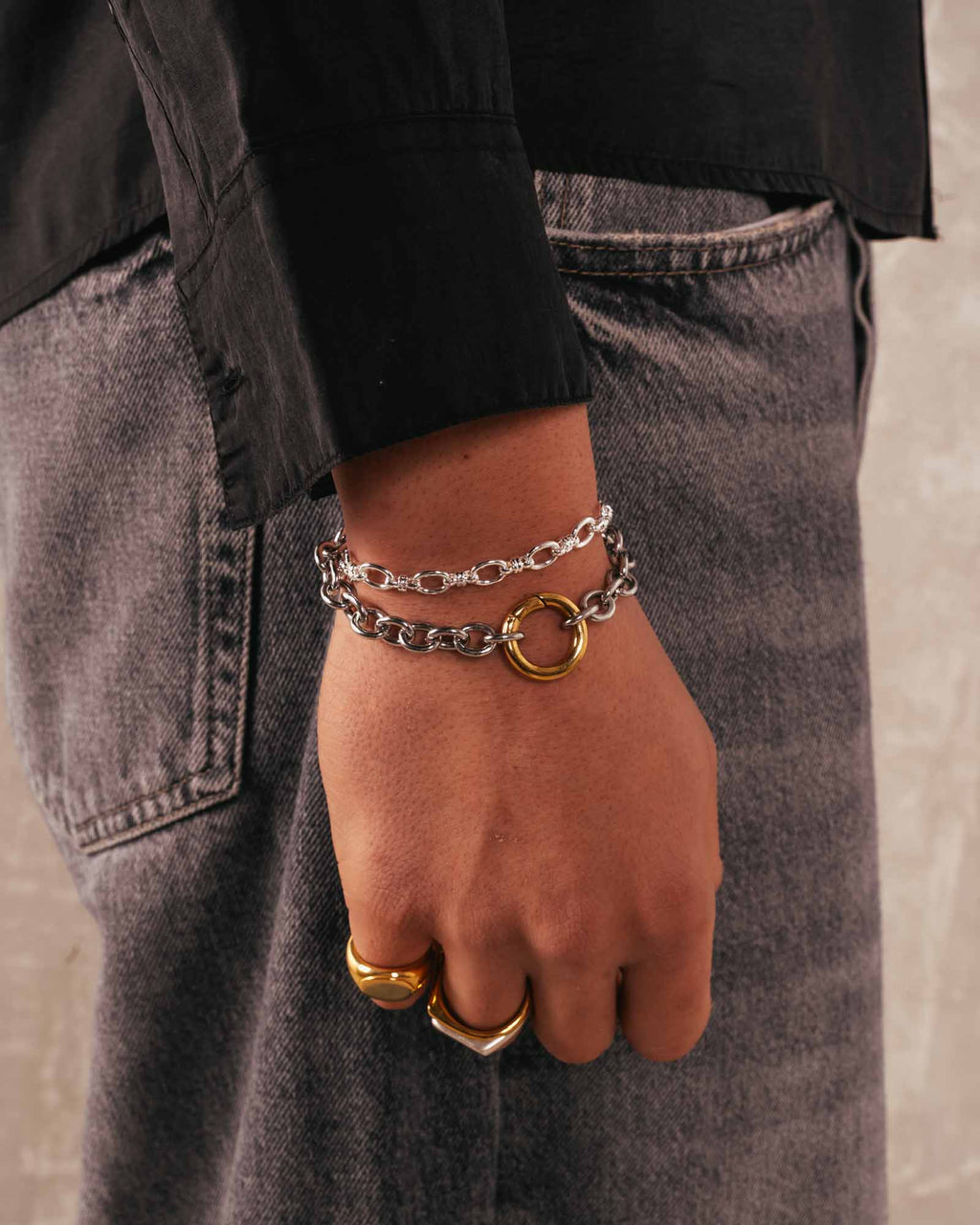 Hand wearing a silver chain bracelet and gold ring on a neutral background