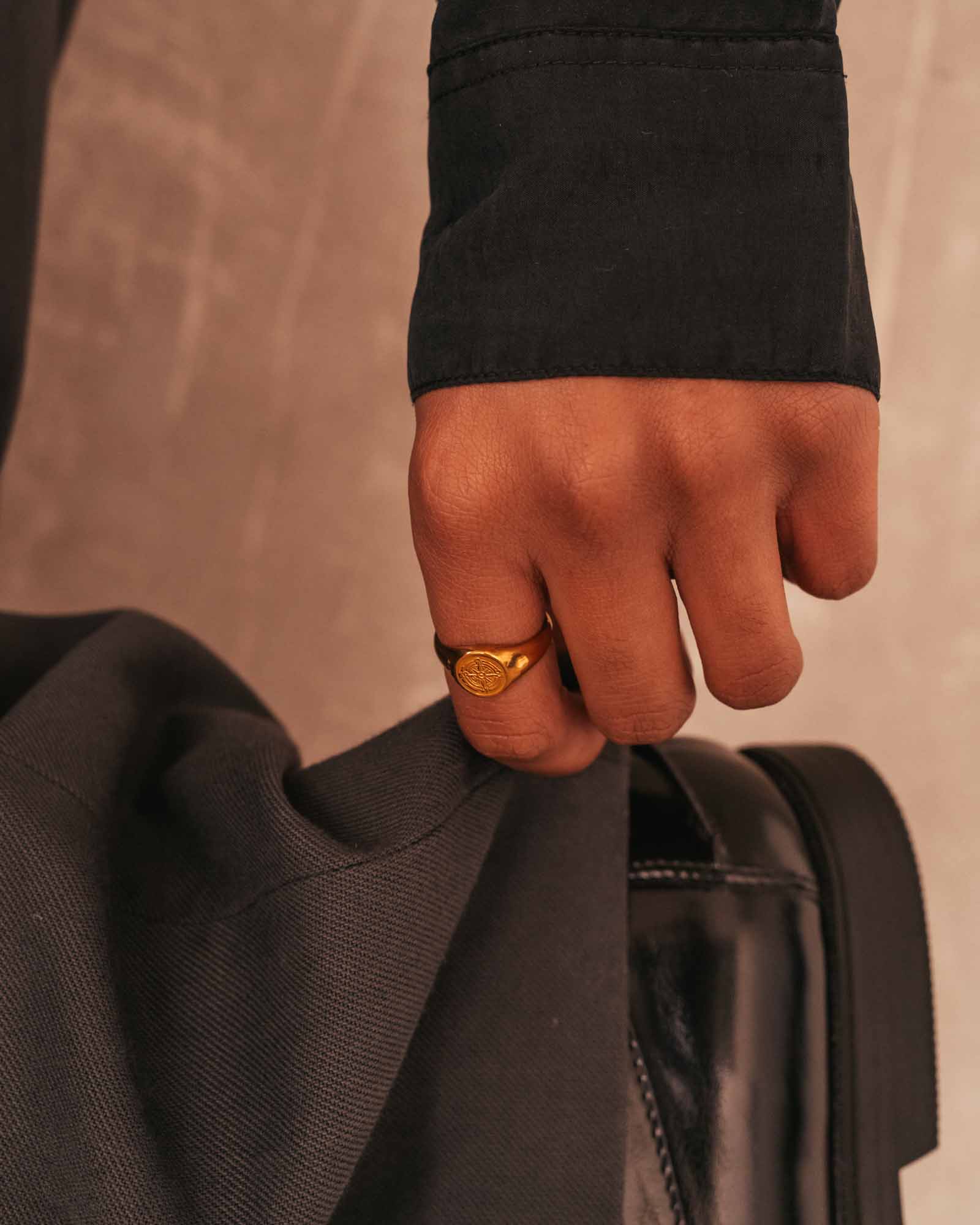 Hand wearing a gold ring holding a black coat against a neutral background