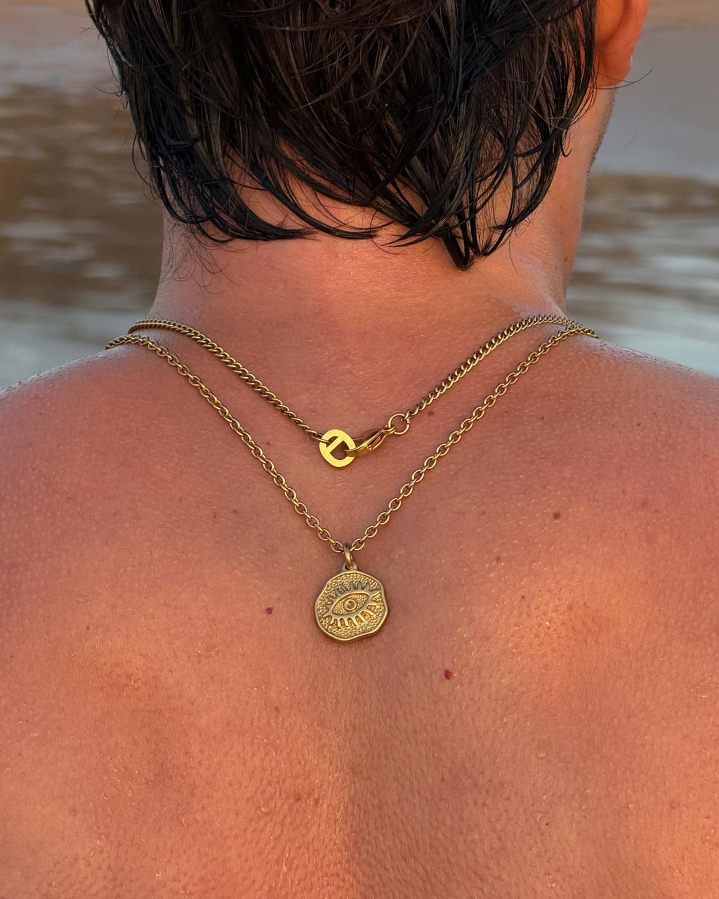 Man wearing gold necklaces with a coin pendant by a body of water.