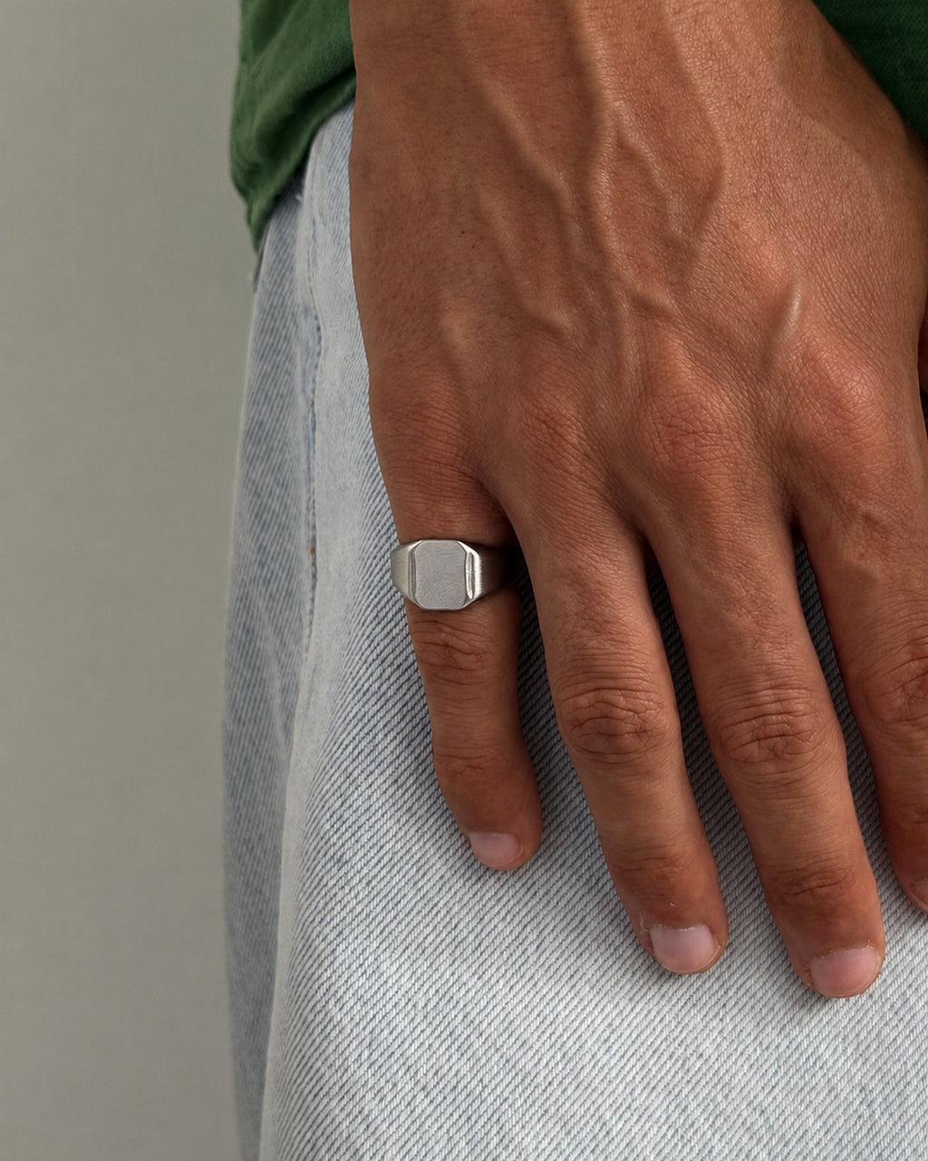 A silver-toned signet ring with a flat, polished rectangular face worn on a person's ring finger, shown against a light gray denim-clad body and a green shirt.