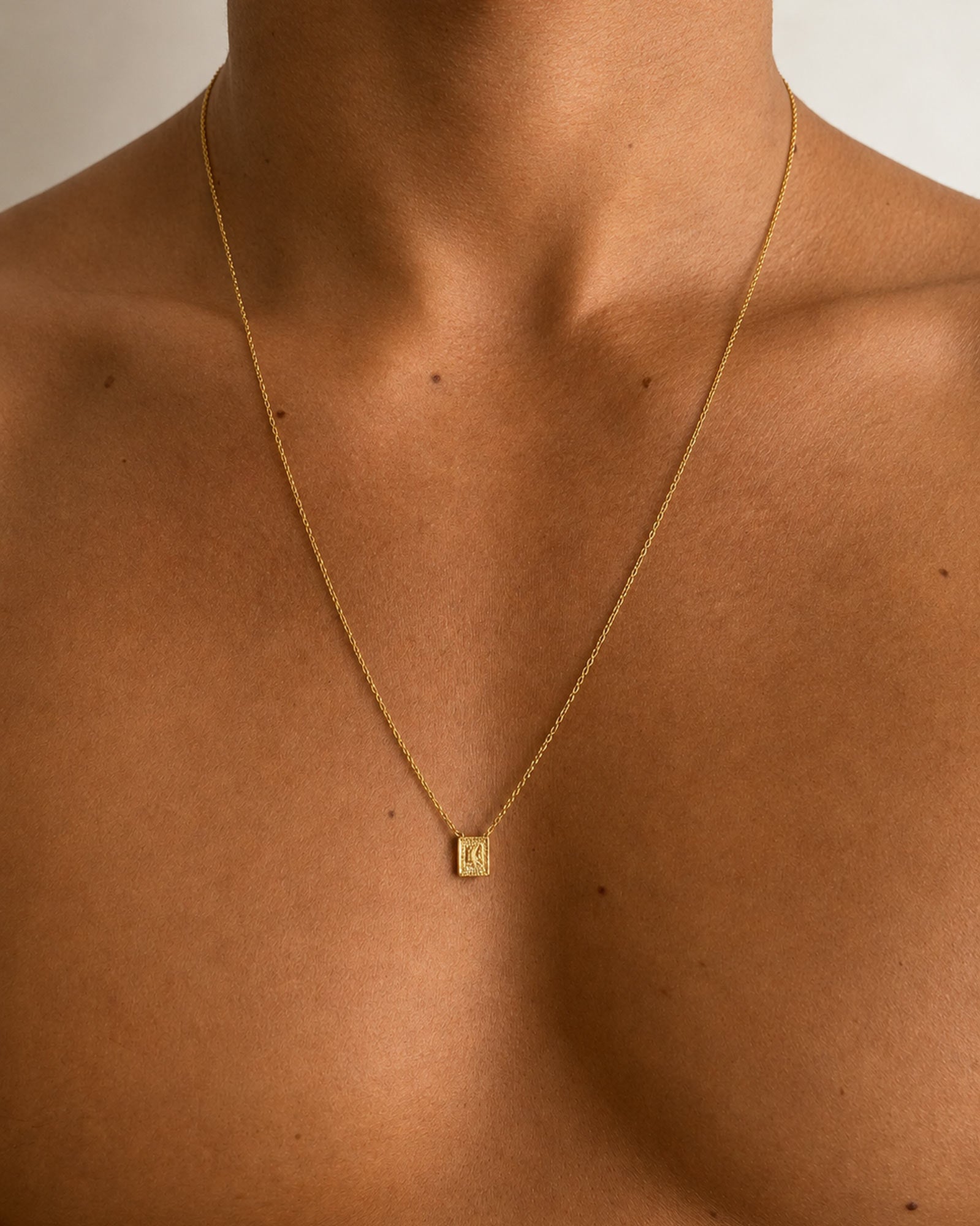 Golden - A close-up of a person's neck and upper chest wearing a delicate gold chain necklace with a small square pendant featuring an engraved letter 'O'.