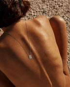 silver - A close-up lifestyle shot of a person's bare back and shoulder, wearing a delicate silver chain necklace with a small rectangular engraved pendant, set against a background of pebbles.