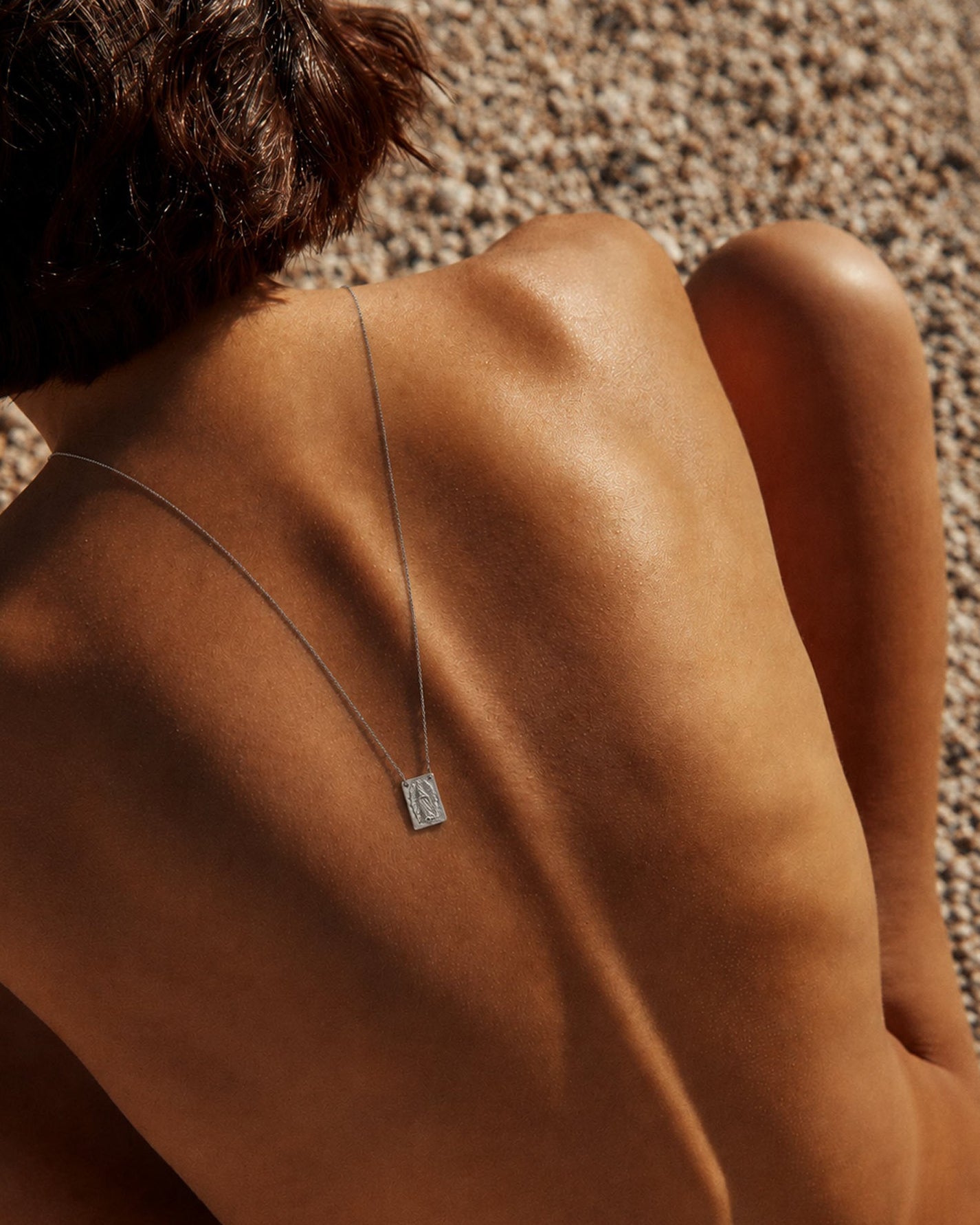 silver - A close-up lifestyle shot of a person's bare back and shoulder, wearing a delicate silver chain necklace with a small rectangular engraved pendant, set against a background of pebbles.