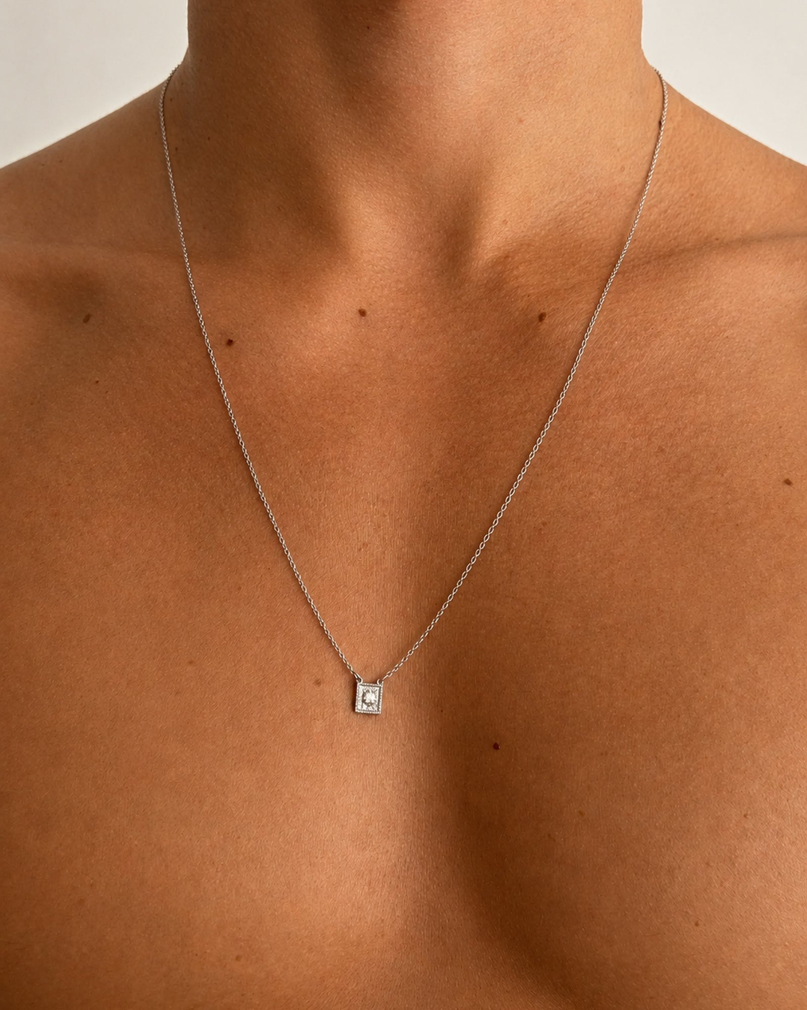 Silver - A close-up of a model's neck and upper chest wearing a delicate silver chain necklace with a small square pendant featuring a central diamond.