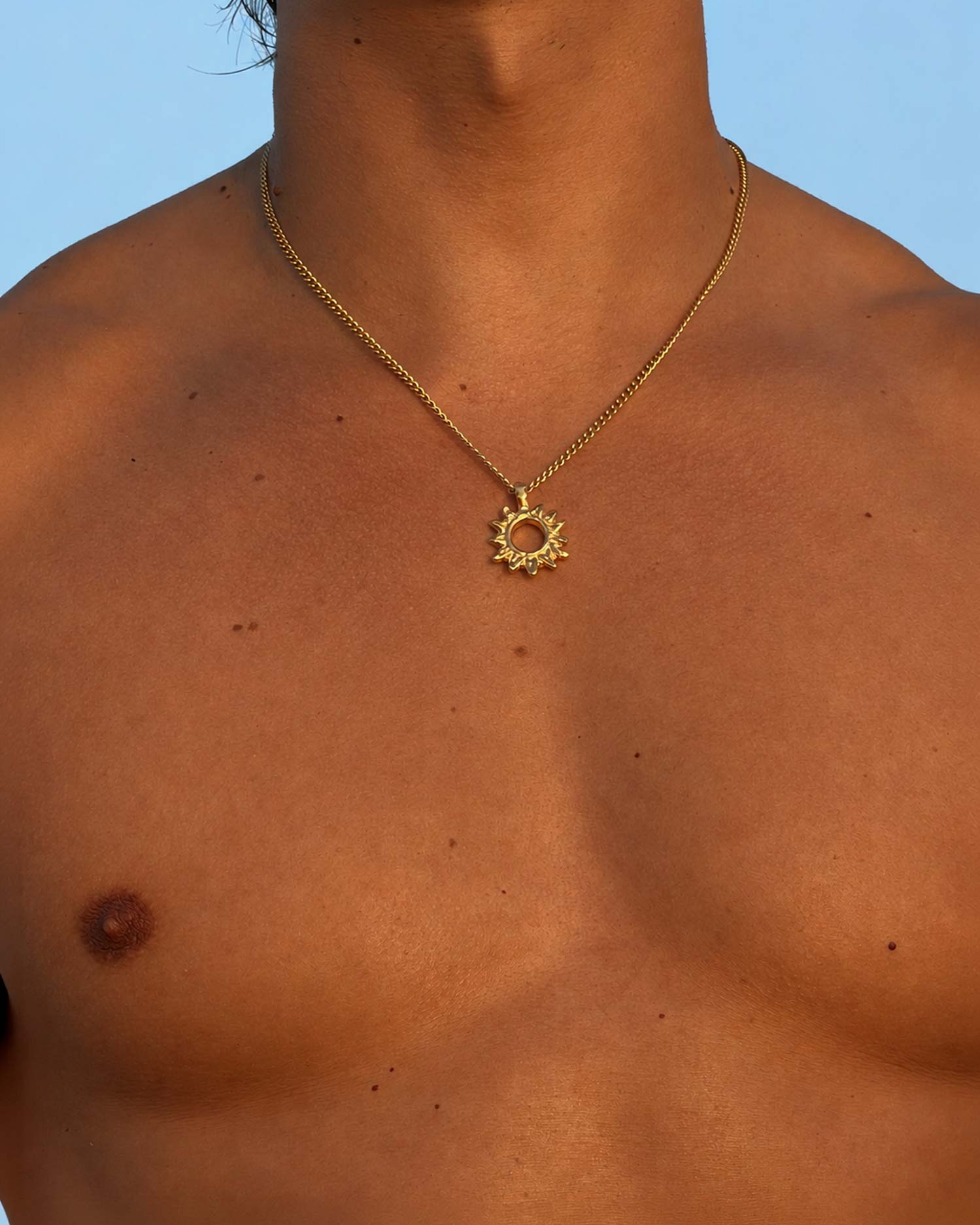 Gold necklace with a sun pendant on a person's chest against a blue sky.