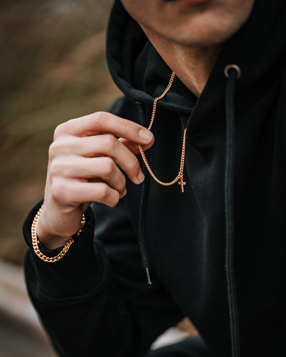 Man wearing black hoodie holding Caprera Golden Necklace with cross pendant and matching bracelet